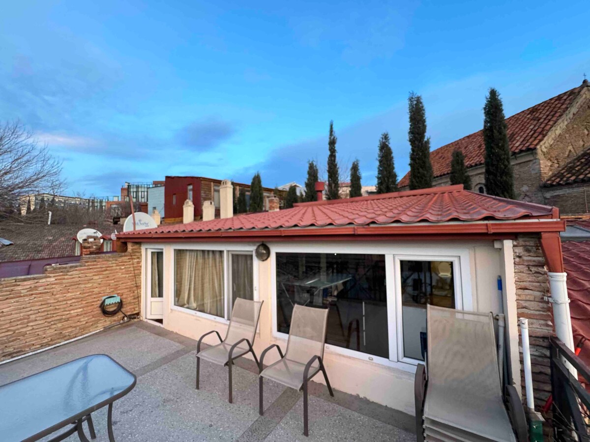 An outdoor terrace is showcased with a spacious area featuring a glass table and several chairs. The roof’s red tiles contrast with the light-colored walls of the adjoining structure. Trees line the background, adding greenery to the view against a bright sky.