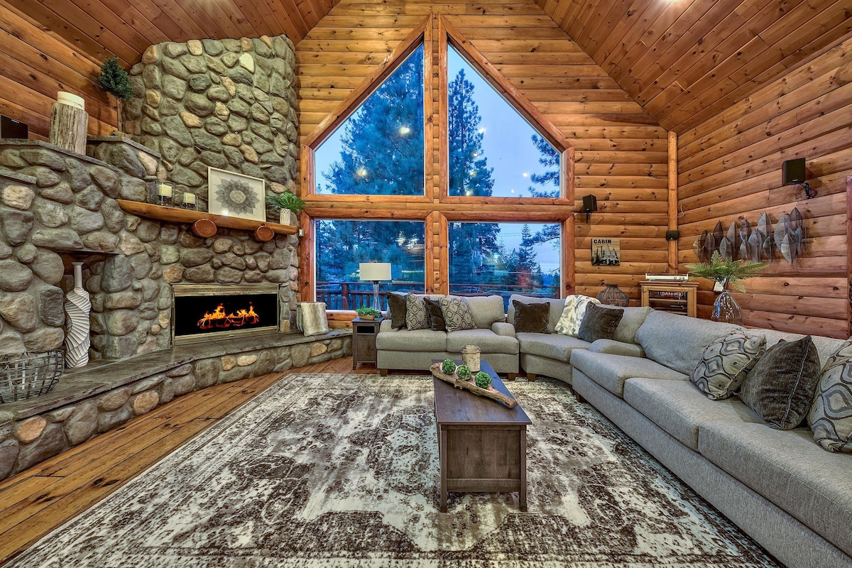 A spacious living area features a large sectional sofa arranged around a coffee table. A stone fireplace is built into the wall, surrounded by wooden accents. Expansive windows provide a view of the outdoors, and a cozy rug enhances the warm atmosphere.