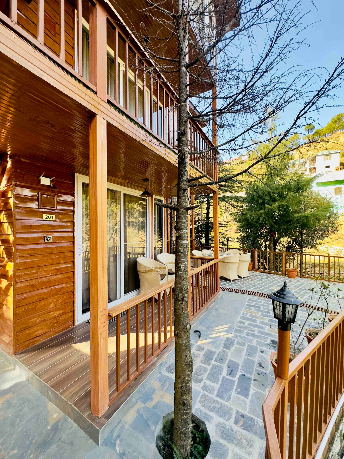 A wooden terrace features a stone pathway and is surrounded by greenery. Two outdoor chairs are positioned on the terrace, providing a relaxing spot. Natural light illuminates the area, highlighting the rich wood tones of the building's exterior.