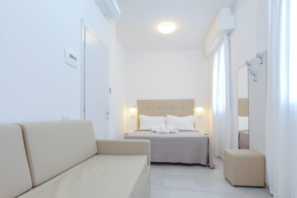 A well-lit guest room features a spacious bed with light bedding, complemented by a soft upholstered headboard. A cream-colored sofa is positioned near the foot of the bed, with a small stool nearby. Natural light filters through white curtains, enhancing the clean and modern aesthetic.
