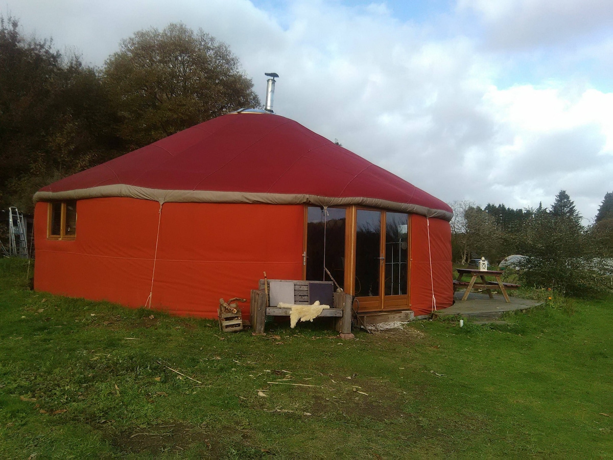 The exterior of a red yurt is shown, featuring large glass doors that provide a view of the surrounding greenery. A wooden deck with seating is visible nearby, complementing the natural landscape. The structure's rounded shape is highlighted against a partly cloudy sky.