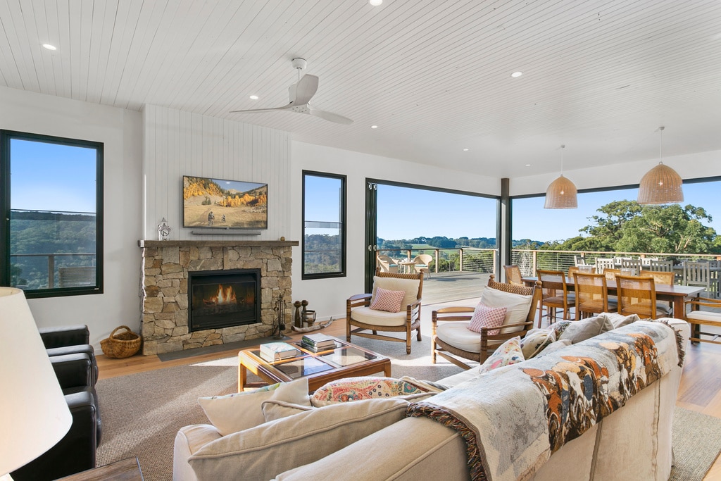 A spacious living area showcases a stone fireplace, surrounded by comfortable seating and natural light from large windows. Wooden accents complement modern furnishings. The open layout leads to a dining area with views of the expansive outdoor deck and rolling hills beyond.