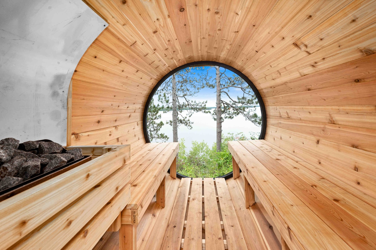 A circular sauna interior, finished with warm wooden planks, features benches on either side. A stone stove sits against one wall. A large circular window offers a view of trees and the lake beyond, allowing natural light to fill the space.