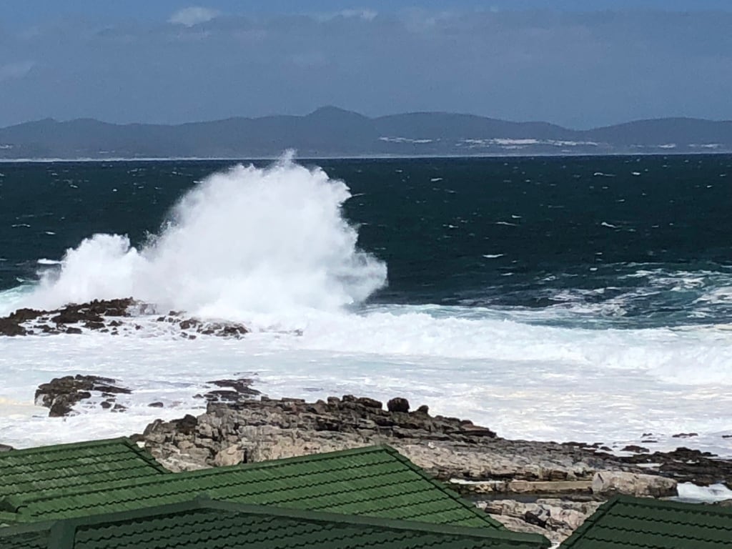 Waves crash against rocky outcrops along the shoreline, creating a spray of white foam. The ocean's deep blue contrasts with the lighter hues of the surf. In the foreground, green roofs of nearby buildings are visible, positioned against the backdrop of the dramatic seascape.