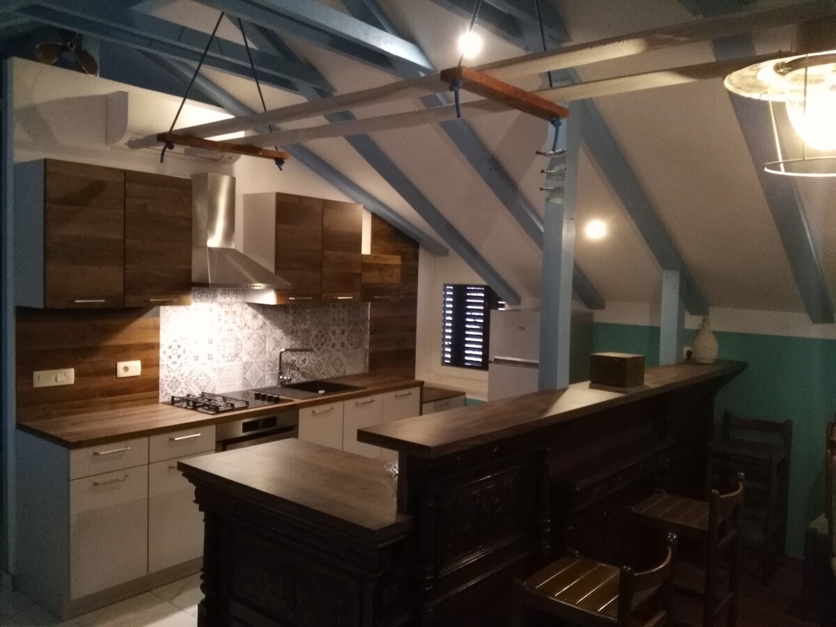 A modern kitchen features wooden cabinetry and a sleek countertop, complemented by a patterned backsplash. Exposed beams highlight the sloped ceiling, while warm lighting offers a cozy ambiance. A bar area with high stools invites social gatherings, enhancing the functional layout.