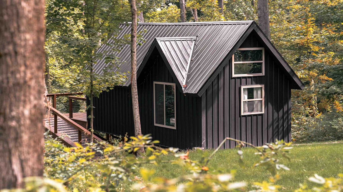 A modern black cabin is surrounded by lush greenery and trees. Its A-frame design features large windows that invite natural light. A wooden deck extends from the cabin, providing a serene connection to the peaceful wooded setting.
