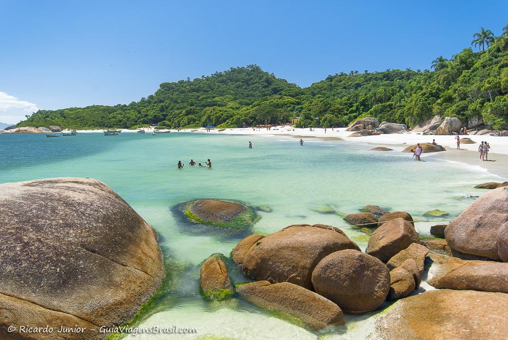 A serene beach scene featuring soft white sands bordered by smooth rocks and clear turquoise waters. Lush greenery rises in the background, while a few individuals can be seen enjoying the water. Gentle waves lap against the shore, creating a calming atmosphere.