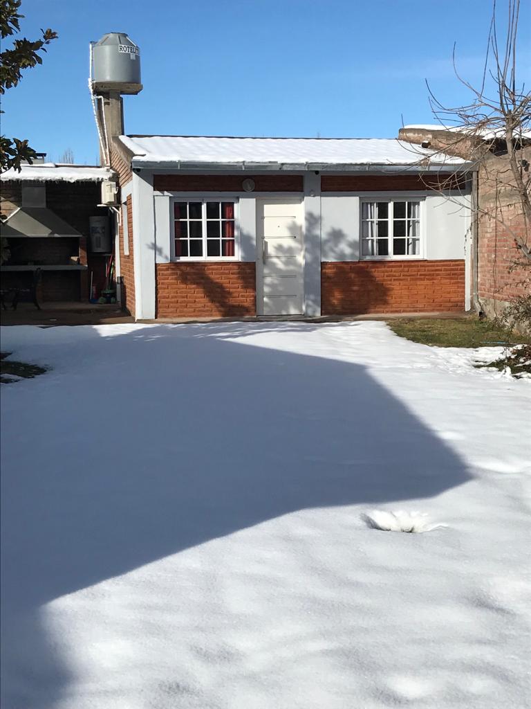 A charming exterior with a snow-covered ground surrounds a single-story house. The front facade features wooden accents and double windows framed in white, allowing sunlight to filter through. A covered area and garden space are visible, contributing to the property's welcoming appearance.