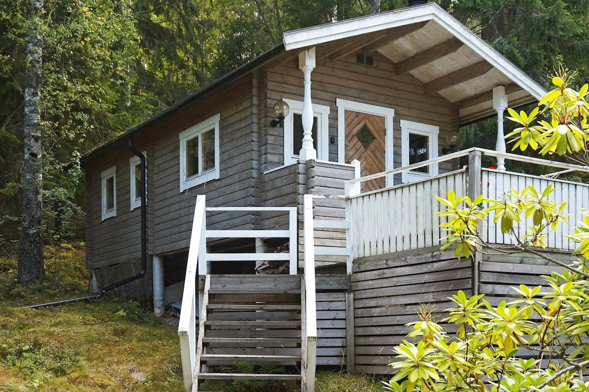 A wooden cottage is seen surrounded by trees, featuring a welcoming terrace with railings. Stairs lead to the entrance, and large windows provide views of the peaceful woodland. The structure is designed with a sloping roof and natural wooden texture, blending with its serene environment.