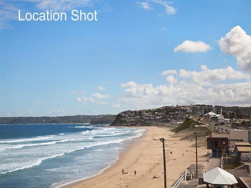 A sandy beach stretches along the coastline, meeting gentle waves under a clear blue sky. The distant hills are dotted with buildings, while the beach area shows a few figures enjoying the sun. The scene captures a serene coastal atmosphere.