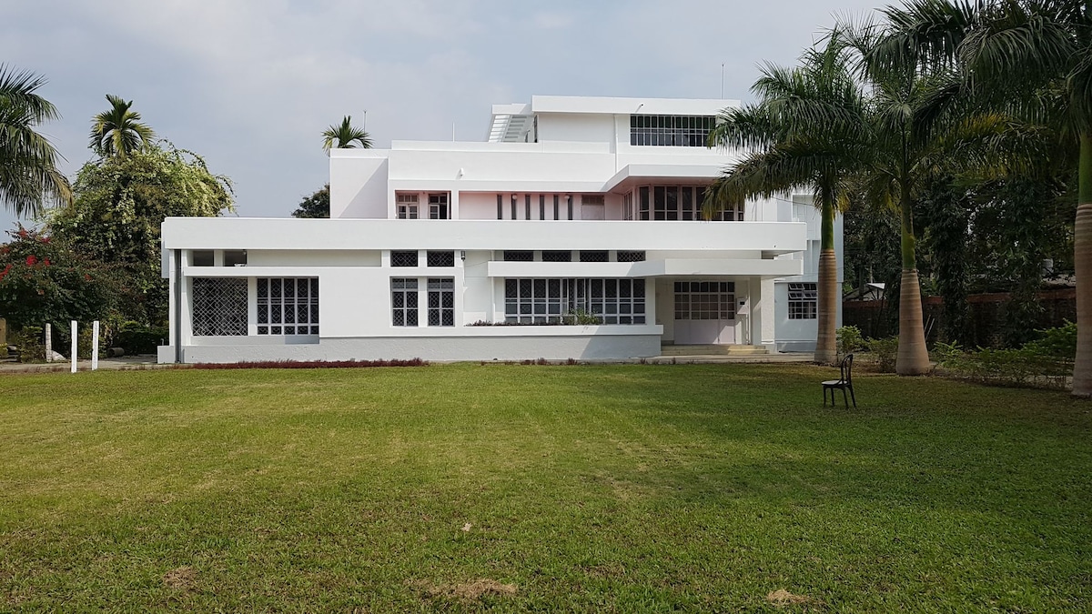 A spacious post-colonial bungalow is set against a clear sky, featuring a white exterior with large windows. The expansive green lawn is complemented by palm trees, providing an inviting outdoor space. A solitary chair is positioned on the grass, adding to the serene atmosphere.