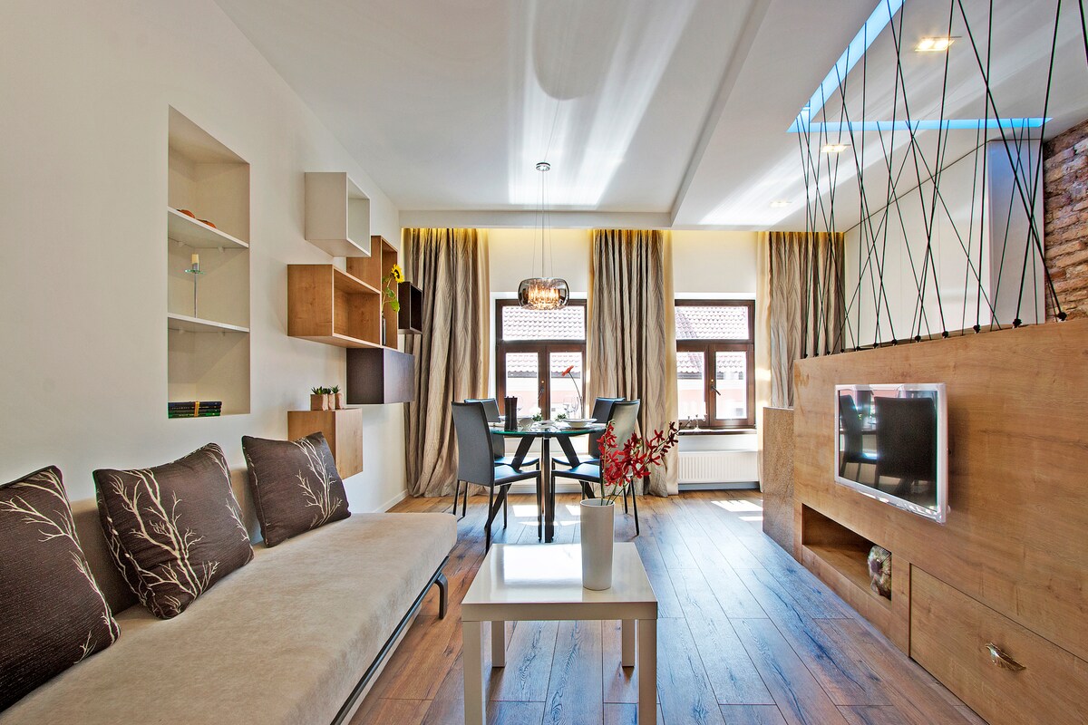 A bright living area features a light-toned wooden floor, large windows with sheer curtains, and a cozy sofa adorned with nature-inspired cushions. A modern dining table is set against the wall, complemented by stylish shelving and a decorative flat-screen TV.