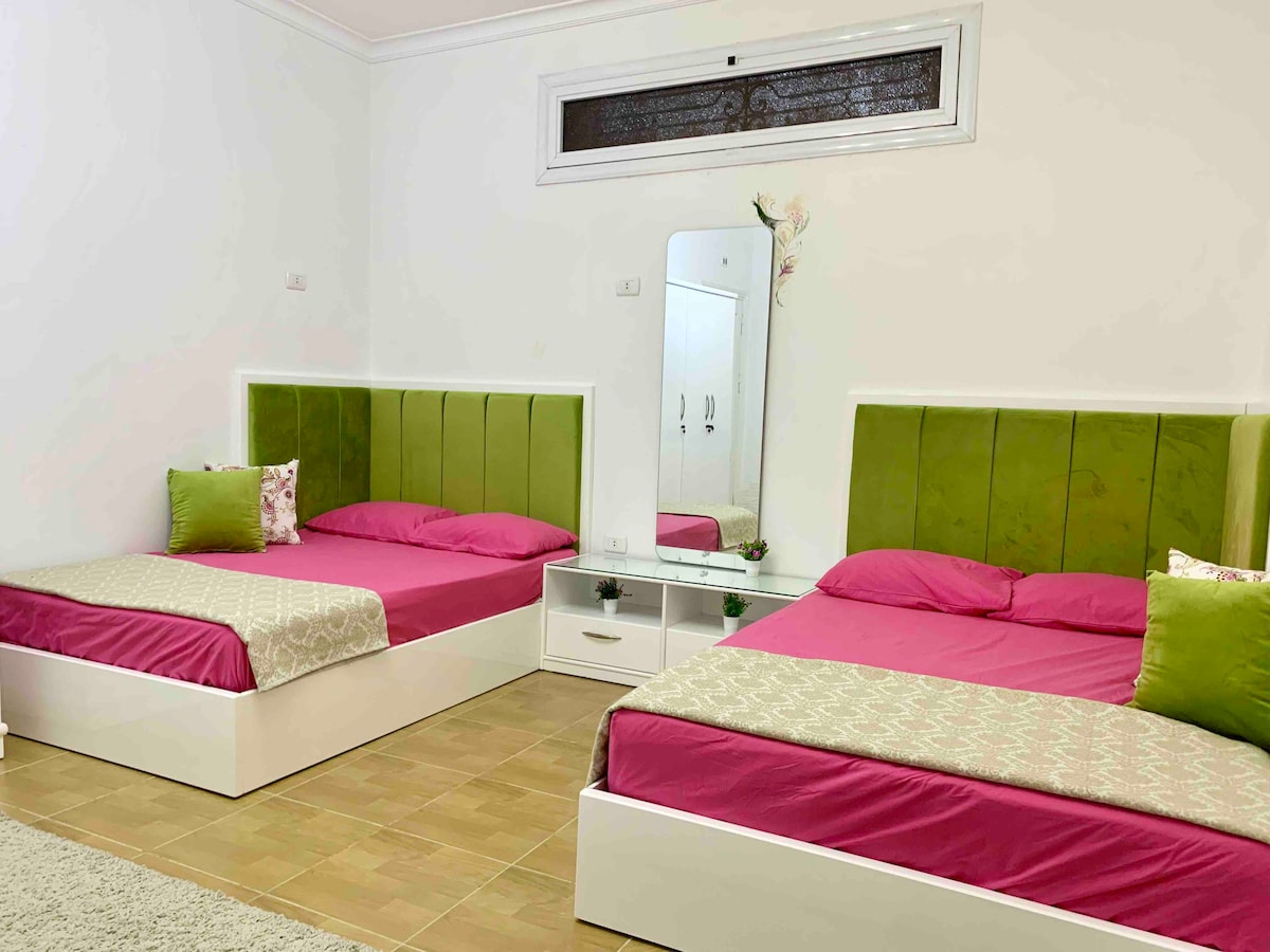 The room features two spacious double beds, each dressed with pink bedding and lime green headboards. A mirrored dresser is positioned between the beds, accompanied by decorative cushions. Soft, beige tiles cover the floor, contributing to a fresh and inviting ambiance.