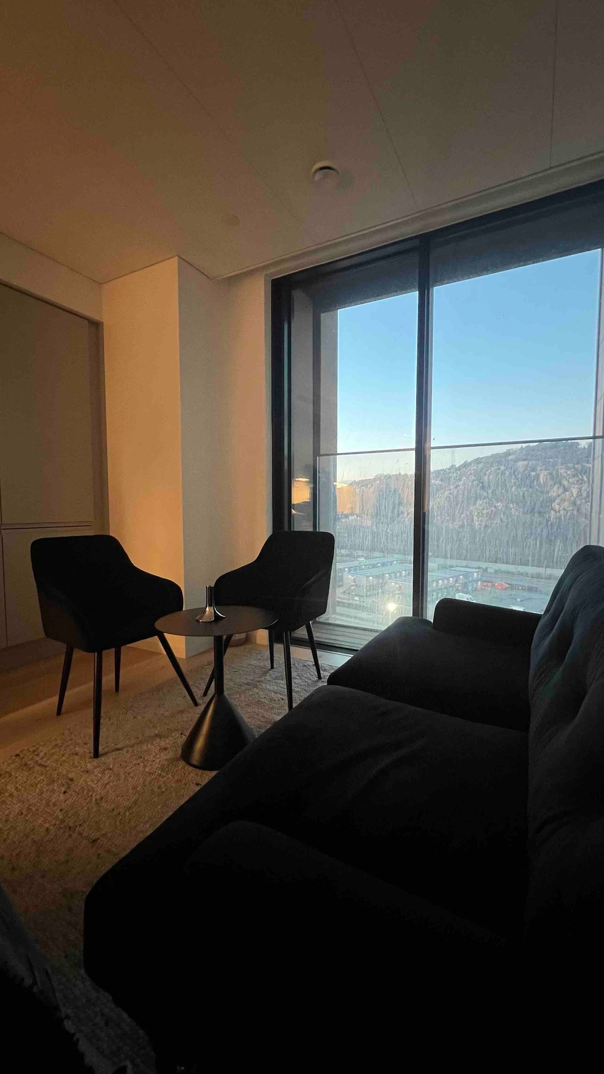 A cozy seating area is highlighted by two black chairs and a small round table. Sunlight filters through the large floor-to-ceiling window, offering a view of the landscape outside. A dark sofa adds comfort to the space within the modern interior.