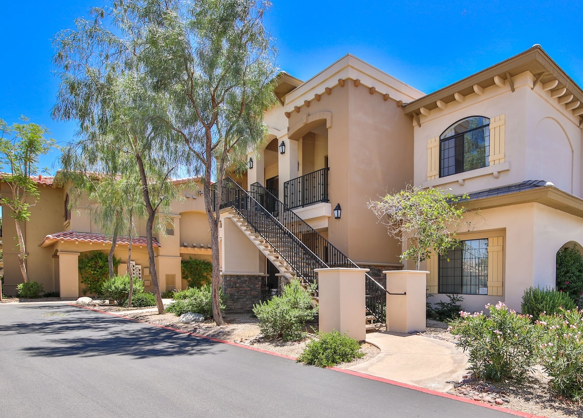 The exterior of a condo complex is presented, featuring a warm beige facade with decorative shutters. A staircase leads to the entrance of the unit, surrounded by lush landscaping that includes trees and shrubs. A paved pathway and driveway provide access to the building.