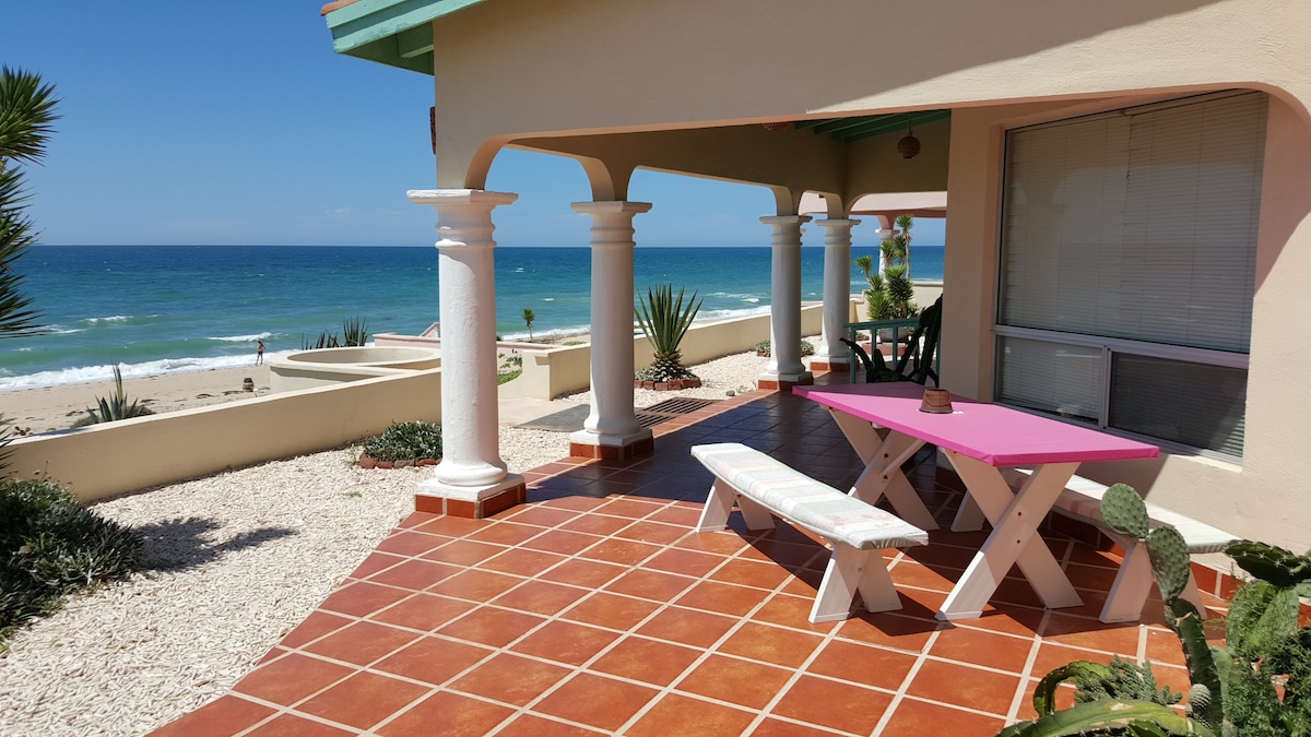 A shaded patio area features a pink table and white bench, surrounded by stone flooring and potted plants. Pillared support frames the view of the sandy beach and ocean, with gentle waves visible in the distance under a clear blue sky.