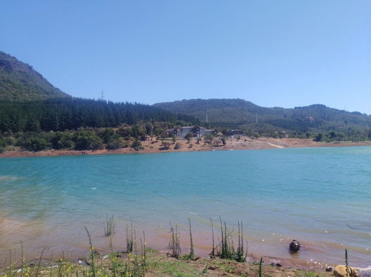 A serene lakeside view showcases a clear, turquoise water surface reflecting a bright sky. The backdrop features rolling hills and native trees, with a settlement visible on the opposite shore, blending harmoniously with the natural landscape.