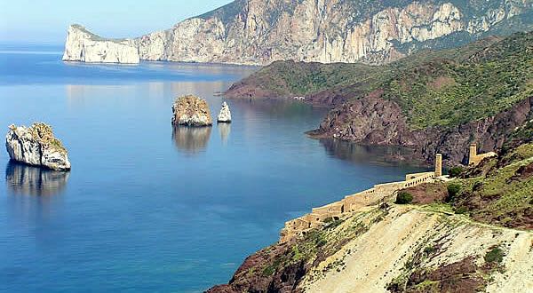 A serene coastal view captures rugged cliffs alongside calm blue waters. Islands protrude from the sea, framed by lush greenery. The coastline is marked by a winding path leading to historical ruins, creating a sense of natural and cultural beauty.