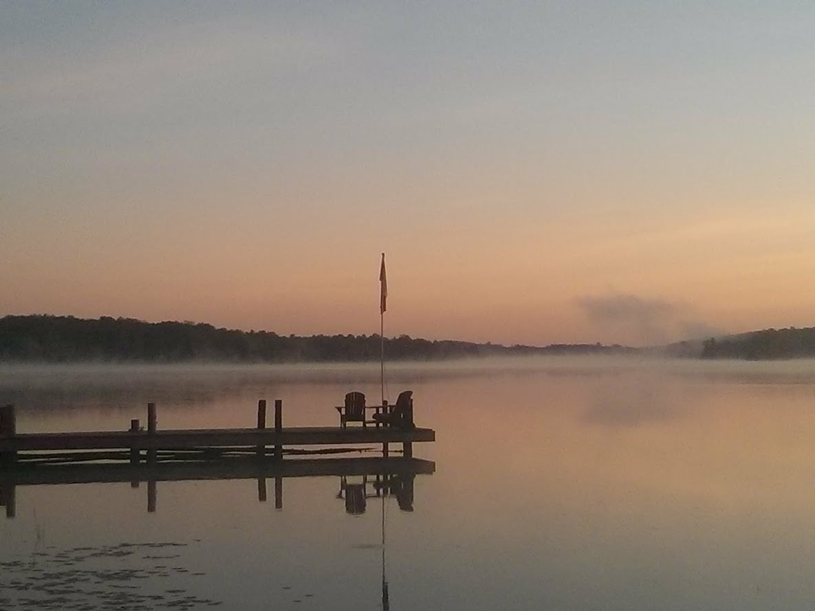 A calm lake is depicted at dawn, with soft colors of orange and pink reflecting on the water. A wooden dock is visible, adorned with an Adirondack chair, and a flag stands tall by the water's edge. Mist rises gently from the lake's surface.