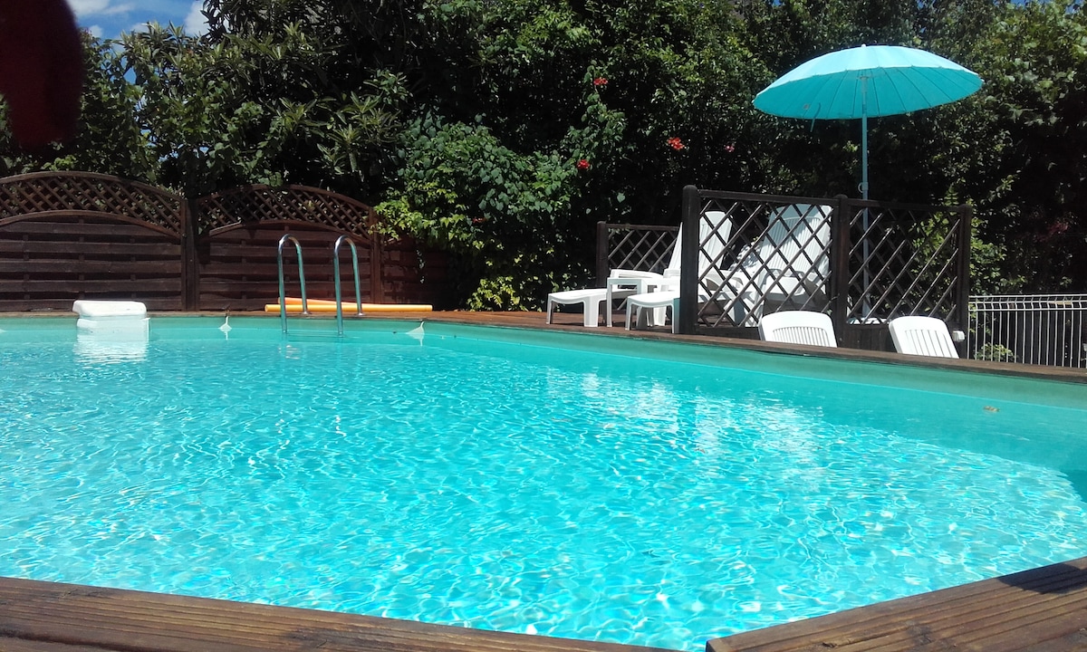 A refreshing pool is surrounded by lush greenery, creating a tranquil setting. An umbrella provides shade over white chairs, while a wooden deck frames the pool area. The clear water is inviting, reflecting the blue sky above, enhancing the overall atmosphere.