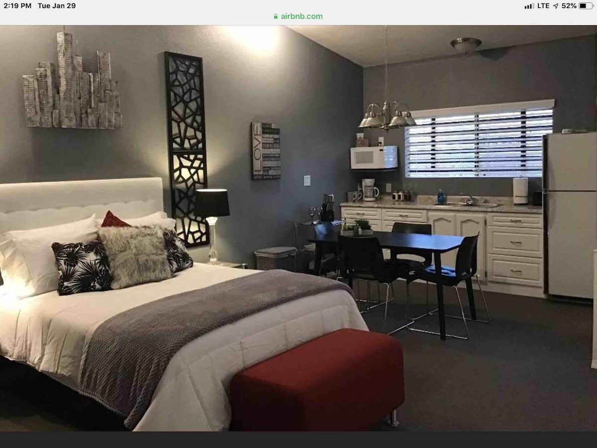 A modern studio features a spacious bed adorned with decorative pillows and a cozy throw, set against grey walls. A small dining area with a table and chairs is visible, alongside a kitchenette equipped with essential appliances, including a microwave and refrigerator.