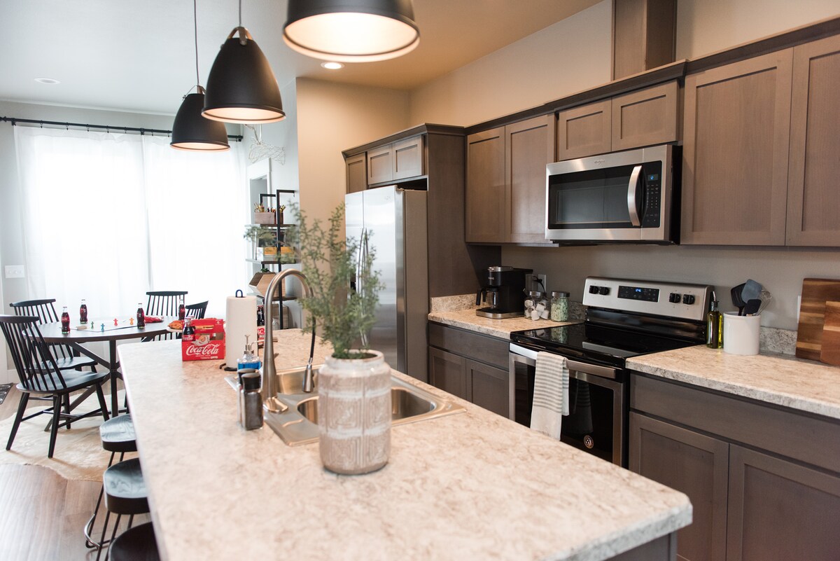 A contemporary kitchen features dark wood cabinetry and stainless steel appliances. A spacious island with a decorative plant at its center is accompanied by a dining area with a wooden table and six black chairs in the background. Natural light filters through large, bright windows.