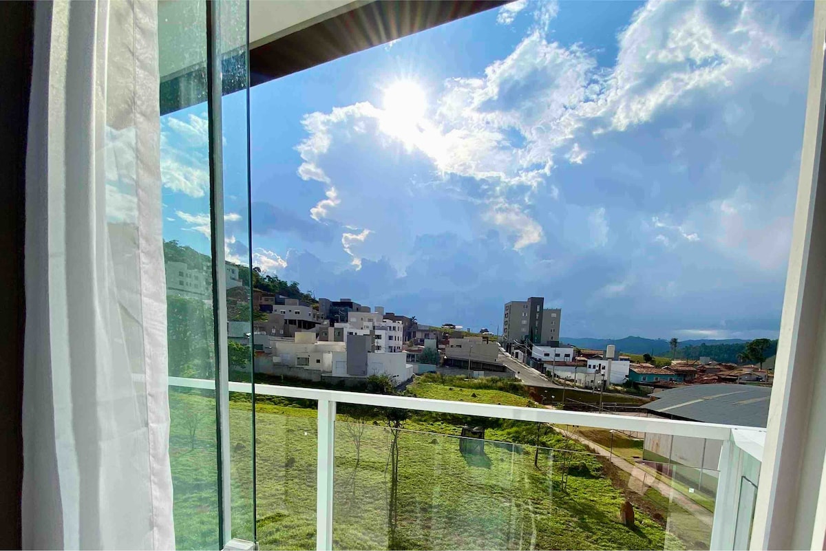 A balcony view showcases a bright sky with scattered clouds and sunlight reflecting off the landscape. Below, residential buildings are visible, surrounded by green fields that extend towards the horizon, providing a serene atmosphere.