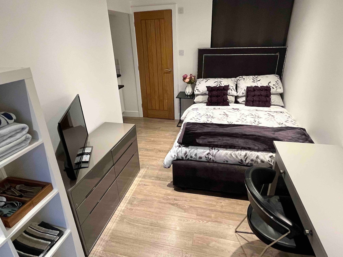 A compact living space is presented with a double bed, dressed in patterned linens. A modern TV is positioned on a dark dresser beside the bed. A small desk and chair are situated against the wall, complementing the warm wood door and light-colored walls.