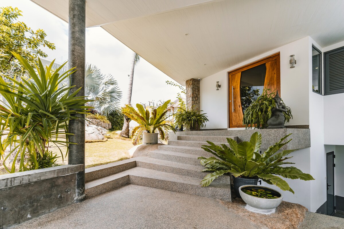 A welcoming entrance features a series of smooth stone steps leading to a large wooden door. Lush green plants are strategically placed around the entryway, creating a serene environment. Natural light filters through, enhancing the peaceful aura of the space.