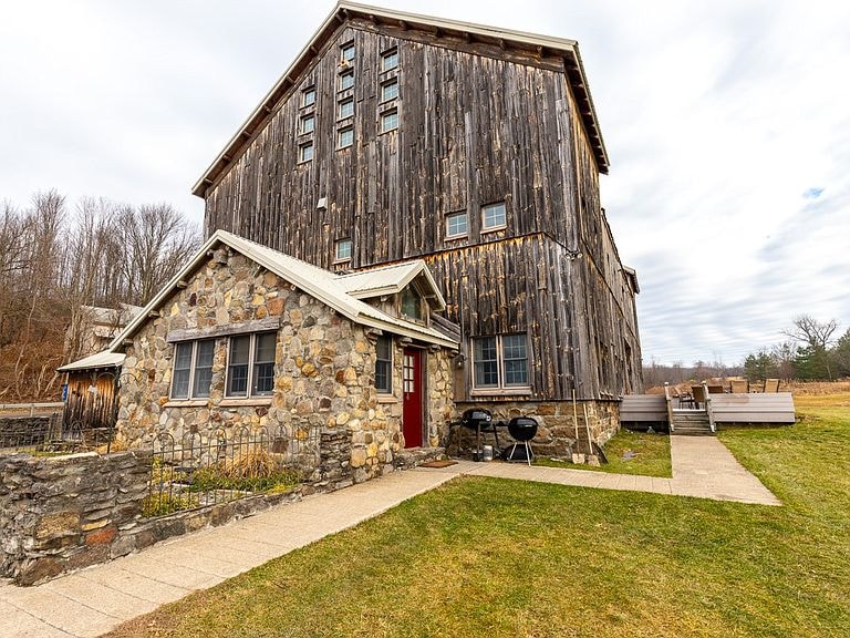 The exterior of the converted barn showcases a blend of weathered wooden siding and stonework. A small patio area features outdoor seating along a paved pathway leading to the backyard, with a barbecue grill positioned nearby. The space is embraced by natural grass and gentle landscape.