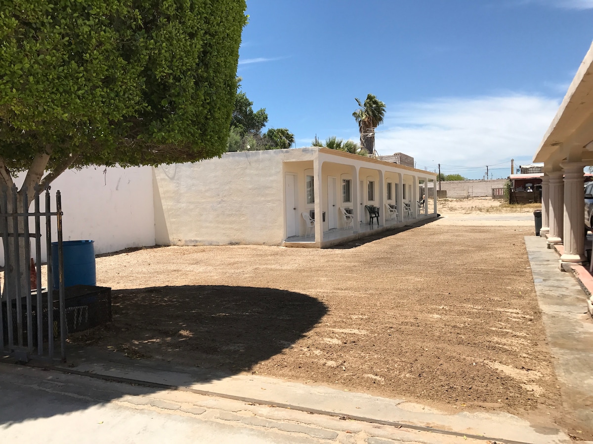 A sandy outdoor area is bordered by a low fence, featuring a row of simple white units with doors, ideal for access. Palms and green foliage are visible in the background, creating a natural environment. The sky is clear with minimal cloud cover.