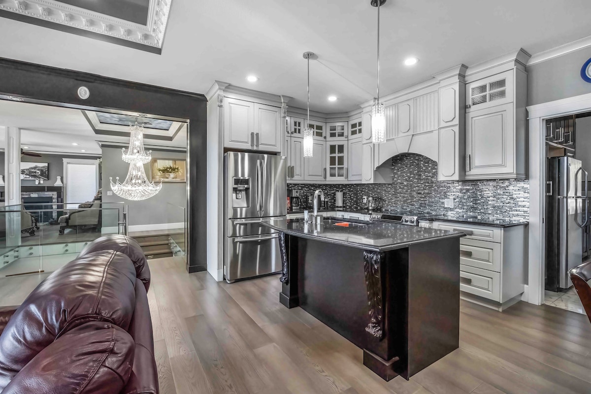 The modern kitchen features sleek cabinetry and a large island with bar seating. Stainless steel appliances, including a refrigerator and gas cooktop, are integrated seamlessly. Pendant lights above the island provide illumination, while an open layout connects the kitchen to the adjacent living area.