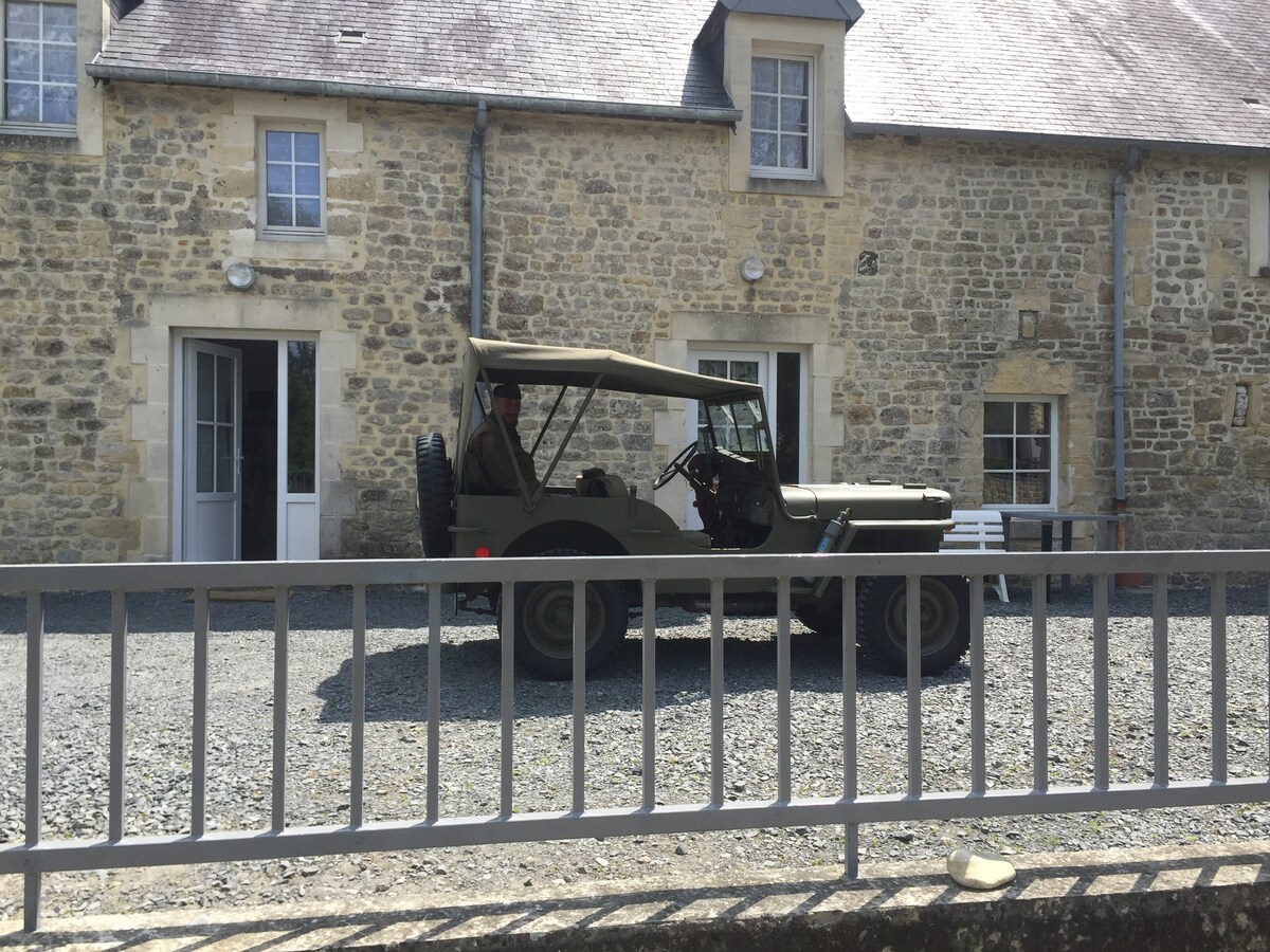 A vintage military jeep is parked on a gravel area in front of a stone building. The building features large windows and a slate roof, blending historical architecture with the surrounding natural environment.