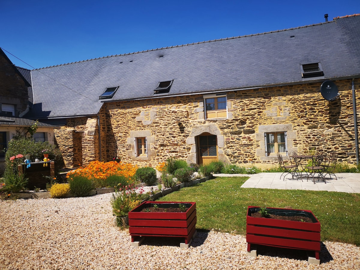 A charming stone facade of a rustic house is shown against a clear blue sky. A well-tended garden features blooming flowers and shrubs, with a patio area containing a table and chairs. Raised garden beds in red wood add a vibrant touch to the outdoor space.