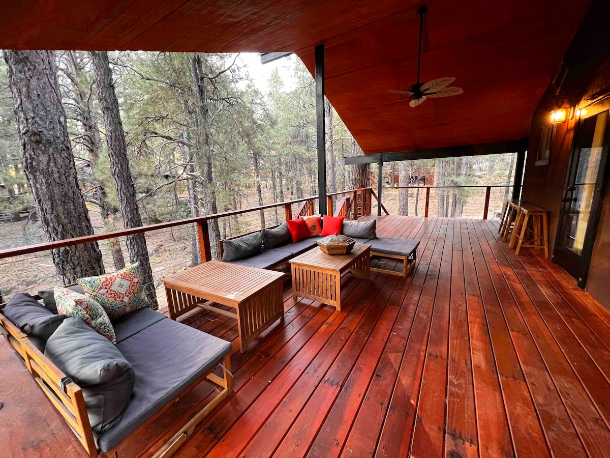 An expansive deck features comfortable seating arrangements, including cushioned couches and a wooden coffee table. The deck, surrounded by tall trees, is designed for relaxation and enjoyment of the serene outdoor environment.