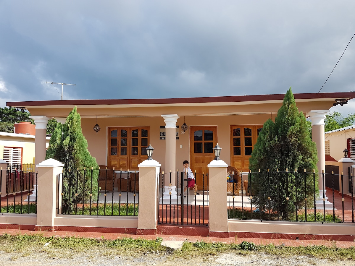 A single-story house is set behind a decorative fence, featuring a light peach exterior and warm wooden doors. Green shrubs flank the entrance, and a covered porch area is visible, providing space for seating. The sky overhead has a mix of clouds, adding depth to the scene.
