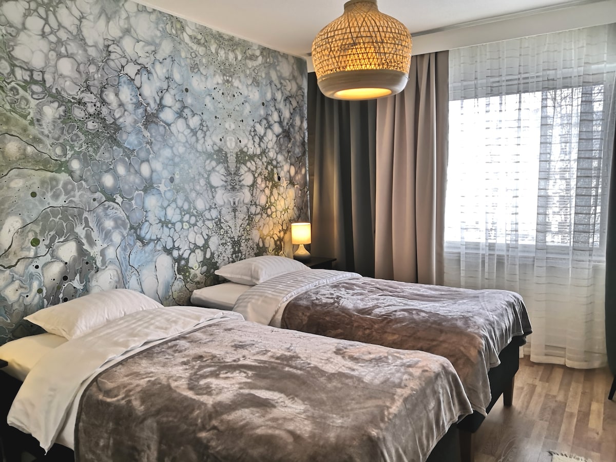 The bedroom features two single beds with plush blankets, positioned next to a softly glowing bedside lamp. A large window allows natural light to fill the room, while a modern, decorative wall mural creates a serene backdrop.