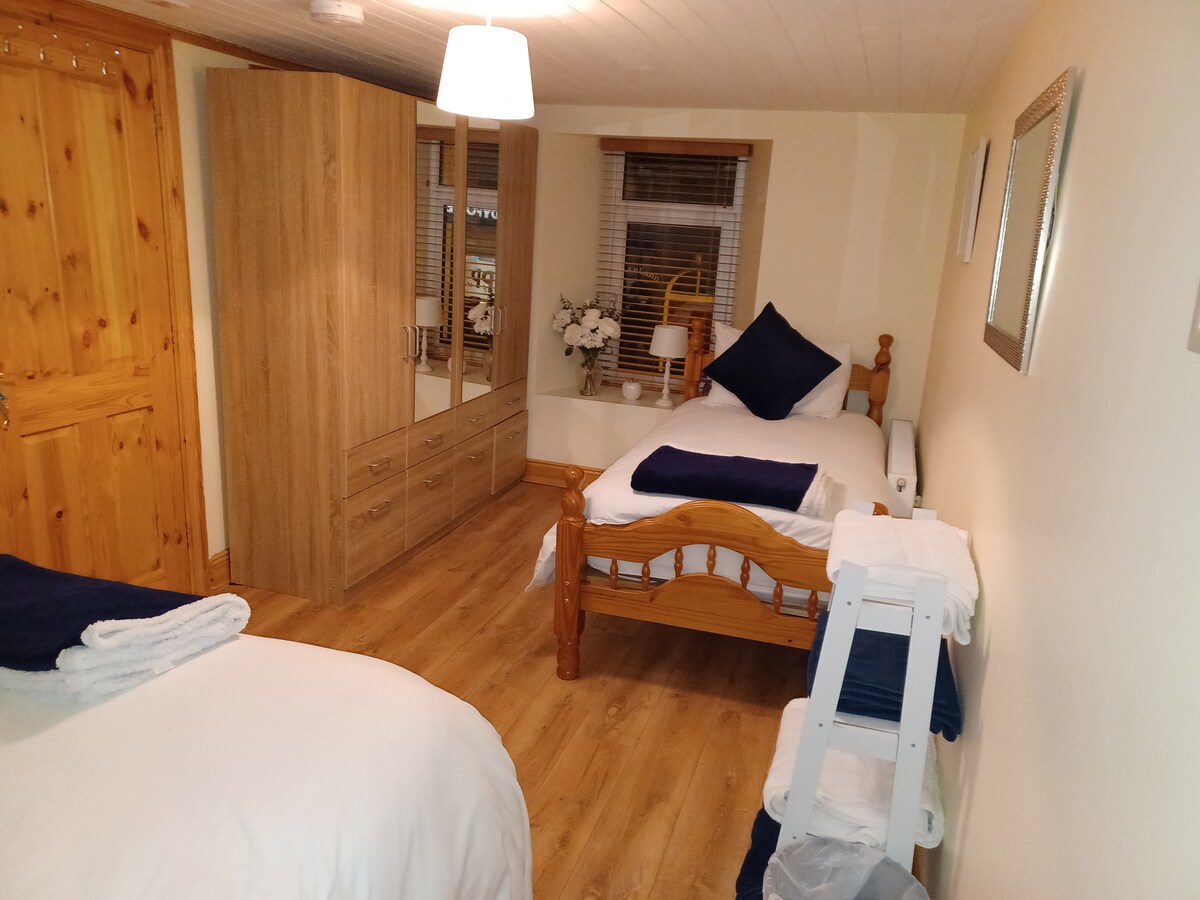 A well-lit room features two beds, one being a spacious double and the other a single, both topped with navy blue cushions. A wooden wardrobe and a mirrored closet provide ample storage. Towels are neatly arranged on a small white rack beside the beds.