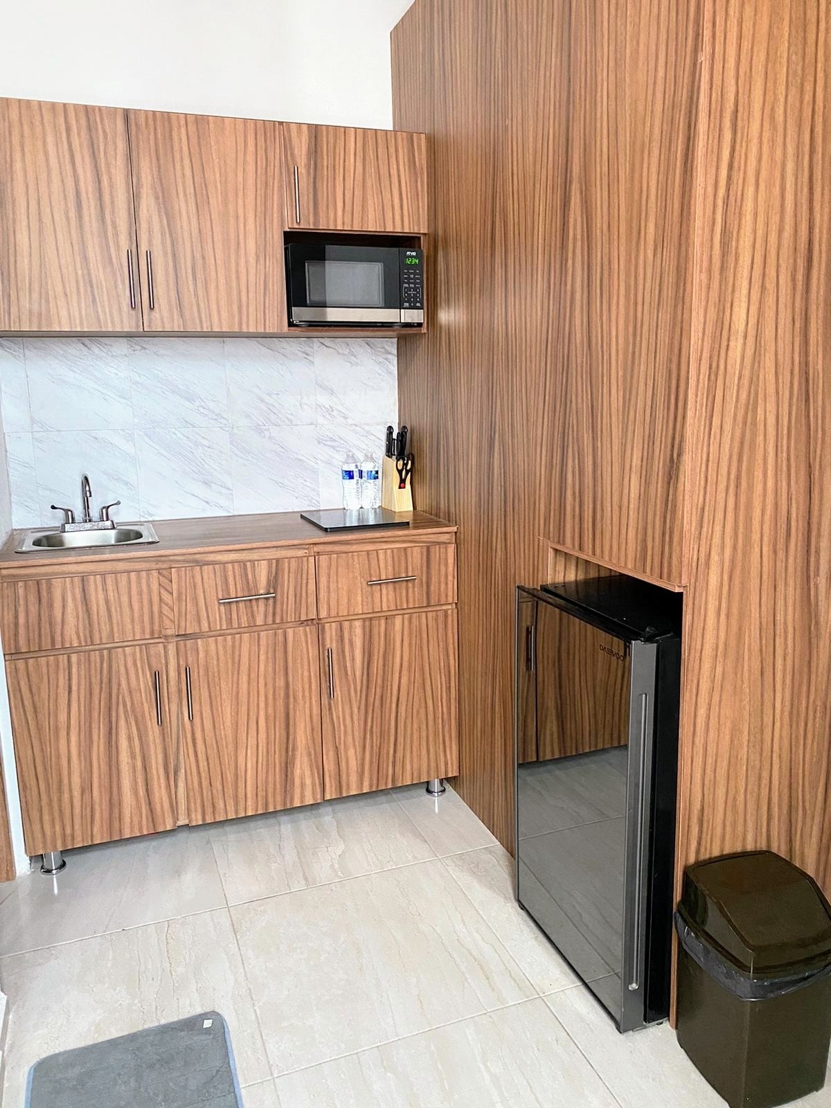 A modern kitchenette is highlighted by warm wood cabinetry. A small sink and a microwave are positioned above a countertop, complemented by utensils in a holder. A compact fridge is located next to a trash bin, and light-colored tiles enhance the space's brightness.