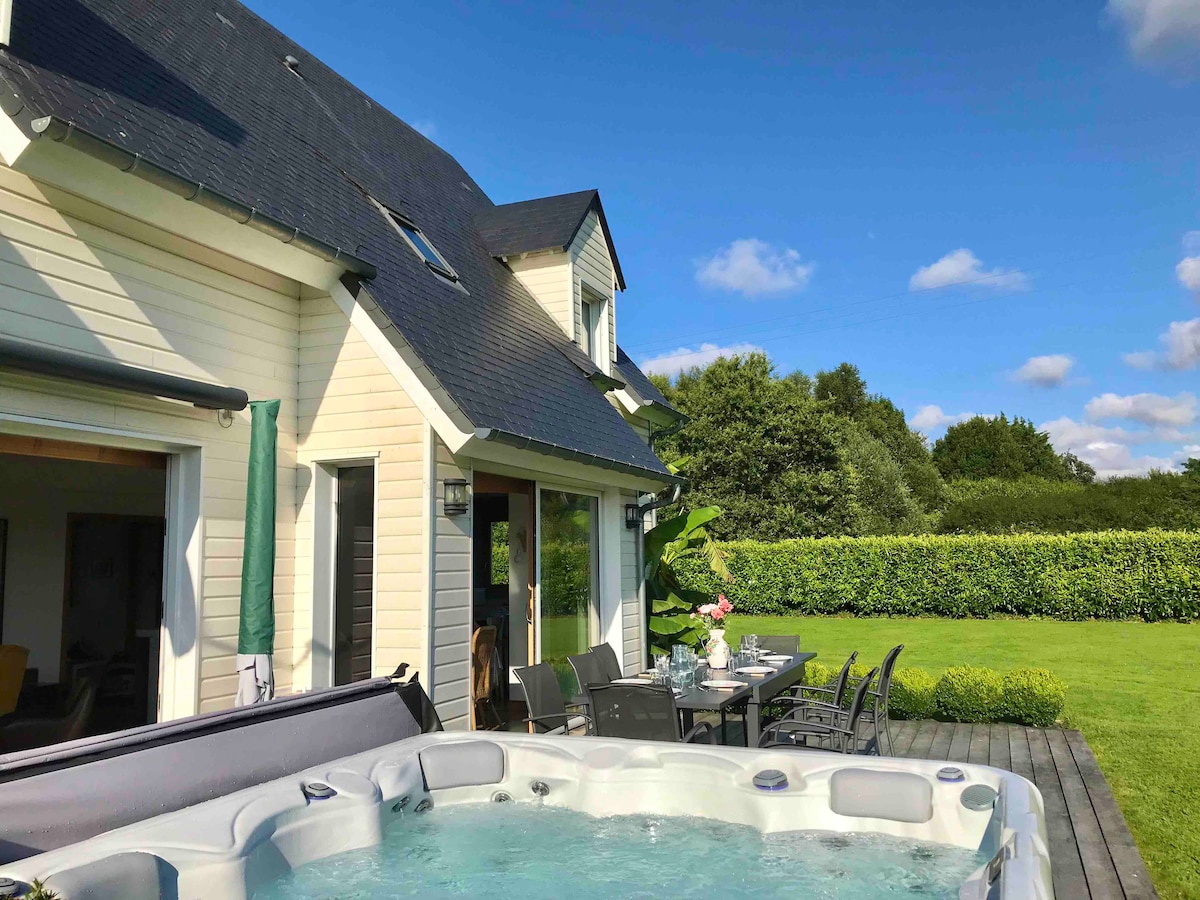 A private terrace features a spacious hot tub, inviting relaxation. Outdoor dining is set for several guests, surrounded by verdant grass and greenery. The exterior of the home showcases light-colored siding and a sloped roof under a clear blue sky.