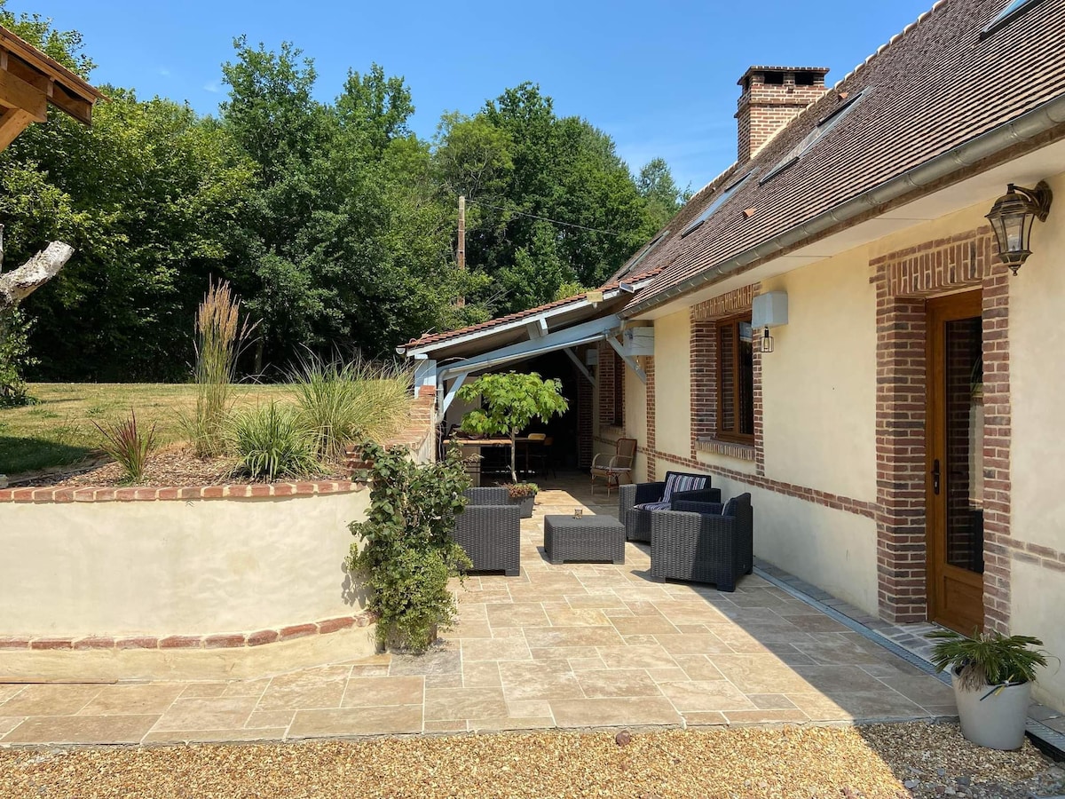 An outdoor seating area is presented with comfortable chairs and a table, surrounded by a stone terrace. Green trees provide shade in the background, while a sloped roof offers shelter over the seating. The combination of brick and light-colored walls contributes to a relaxed atmosphere.