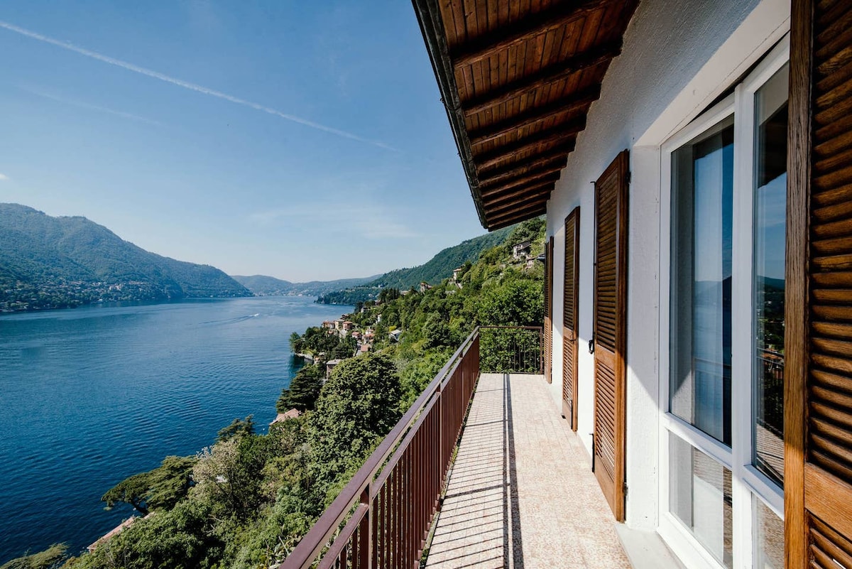 A spacious terrace extends from the villa, offering panoramic views of Lake Como and surrounding mountains. Large windows are flanked by wooden shutters, allowing natural light to fill the space. The surface of the terrace is light-colored, complementing the greenery in the background.