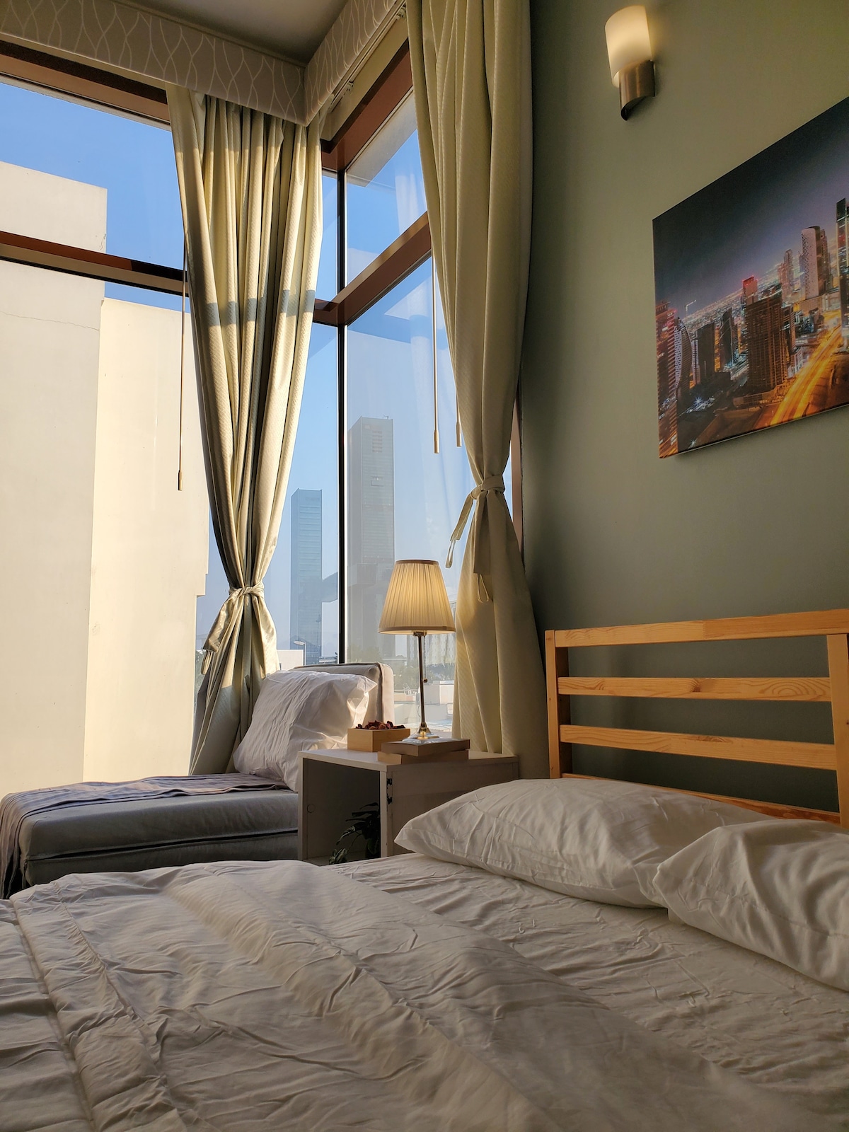 A cozy sleeping area features a queen bed with crisp white linens, complemented by a soft sofa bed nearby. Large windows allow natural light to filter in, providing a glimpse of the Dubai skyline. A bedside lamp offers warm illumination during the evening.
