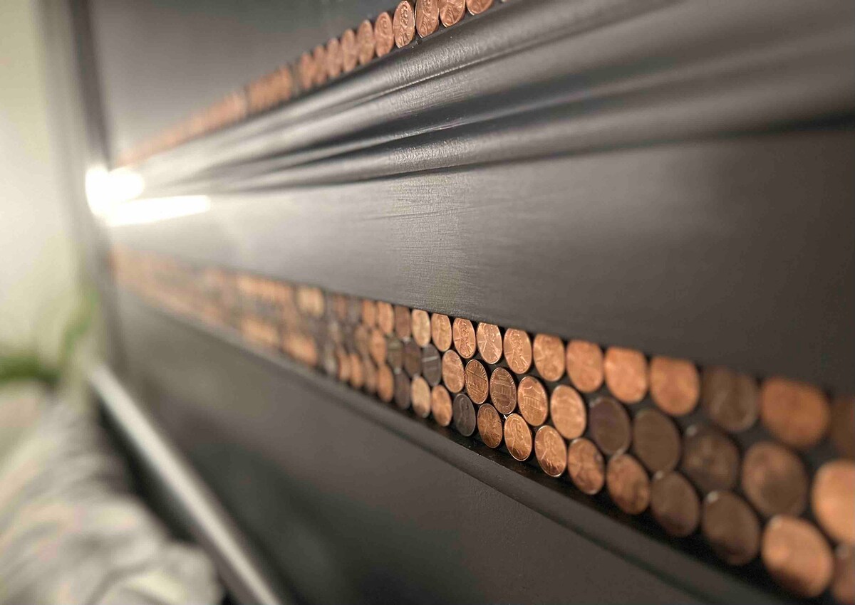 A unique headboard features a decorative band made of closely arranged pennies, set against a sleek, dark-colored surface. The arrangement creates a visually interesting contrast, inviting attention to the craftsmanship. Soft lighting highlights the shiny surfaces of the coins.