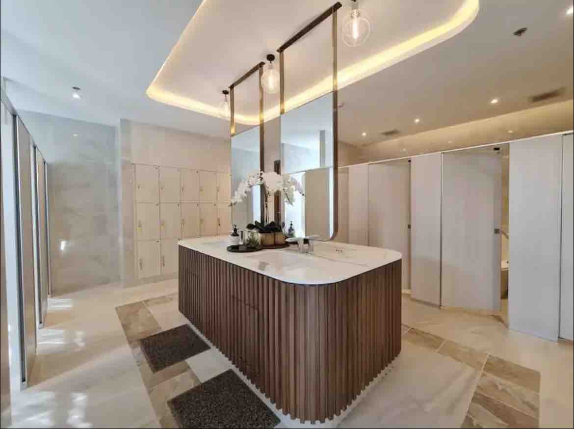 A spacious bathroom area features a large wooden vanity with a white countertop, surrounded by elegant pendant lighting. Locker units line the background, while modern restroom stalls are positioned on the side, completing the contemporary design.