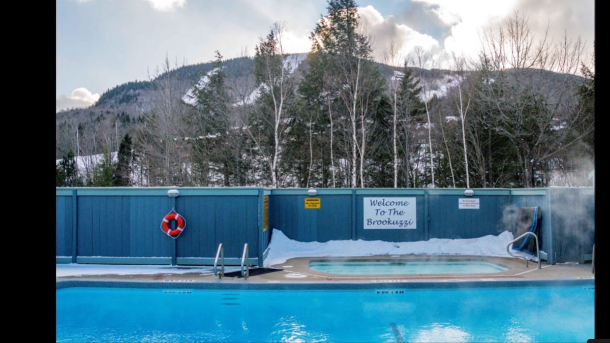 An outdoor pool area is seen, surrounded by snow-covered ground and trees. A hot tub is situated beside the pool, with steam rising into the cool air. A sign that reads 'Welcome To The Brookeggs' is visible on the fence. Mountains are in the background.