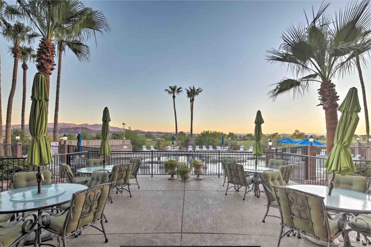 A patio area is presented, featuring several tables and chairs surrounded by lush palm trees. Umbrellas provide shade over the seating, and a view of a serene landscape is visible in the background, including gentle hills and blue skies.