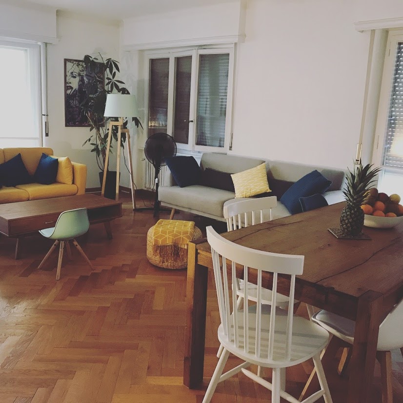 A spacious living area features a yellow sofa paired with a gray couch adorned with blue cushions. A wooden dining table with white chairs sits beside a low coffee table. Natural light flows through the large windows, highlighting the warm wooden flooring.