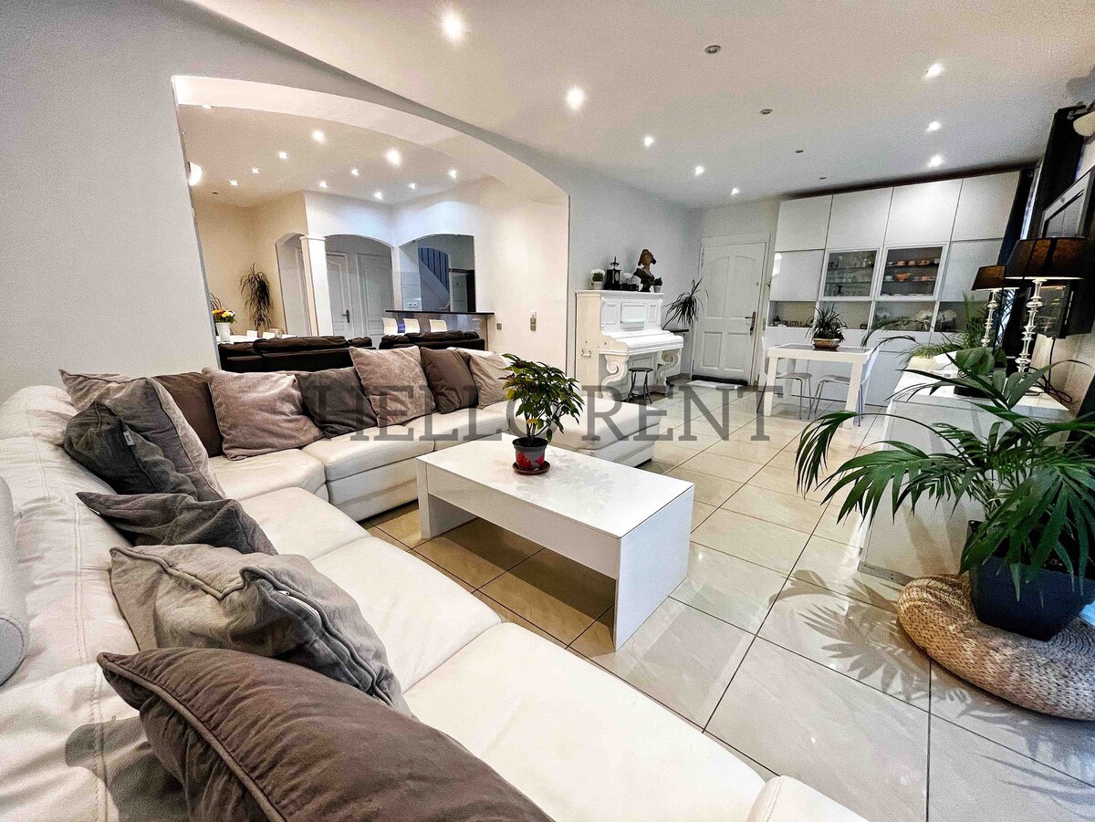 A spacious living area features a large sectional sofa adorned with cushions, arranged around a sleek coffee table. Natural light is provided by recessed ceiling lights and downlights, illuminating the light-colored tiled floor. A potted plant adds a touch of greenery to the contemporary design.