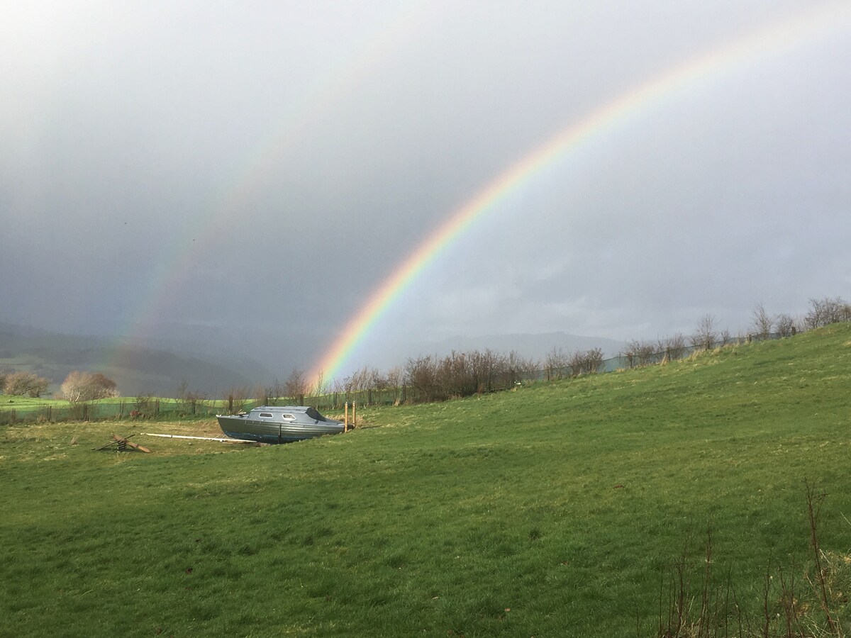 A 1960s wooden yacht is positioned on a grassy landscape, beneath a vibrant double rainbow stretching across the sky. Rolling hills and scattered trees form the backdrop, creating a serene natural setting.